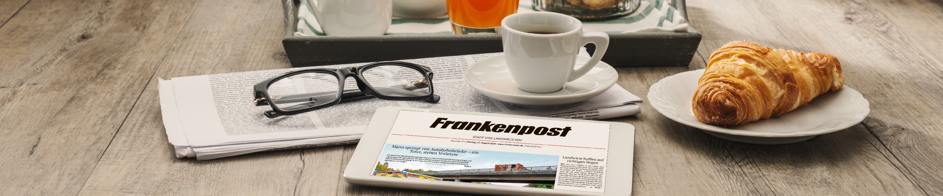 Tablet displaying a newspaper app on a wooden table, with a croissant, cup of coffee, and reading glasses nearby on a tray.